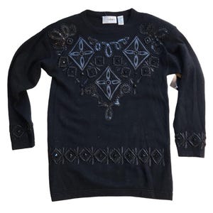 Może przedstawiać: Czarny sweter z długim rękawem z misternymi zdobieniami z koralików. Wzór zawiera geometryczne i kwiatowe wzory w kolorze czarnym i srebrnym. Sweter ma okrągły dekolt i koralikowe detale na mankietach i obszyciu. Widoczna jest nazwa marki "Cedari".