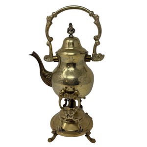 May include: Antique brass samovar with ornate detailing. The tea kettle features a curved spout, a decorative handle, and a small figure on the lid. The samovar sits on a round base with three legs, showcasing an aged, vintage aesthetic.