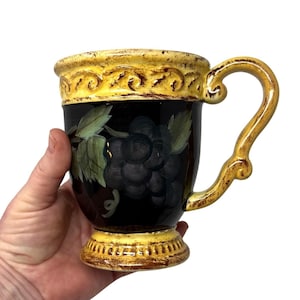 May include: A decorative ceramic mug with a black background and a painted grape design. The mug has a yellow rim and handle, and a decorative base. The mug is held in a hand.