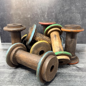 Puede incluir: Una colección de bobinas de madera vintage con cuerpos cilíndricos y bridas circulares. Varias bobinas tienen bordes pintados en colores que incluyen azul claro, amarillo, verde y naranja, mientras que otras mantienen un acabado de madera marrón oscuro.