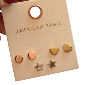 May include: A set of stud earrings on a light beige card. The earrings include two rose gold circles, two gold hearts, and two silver stars. The card is branded with the text "AMERICAN EAGLE" at the top.