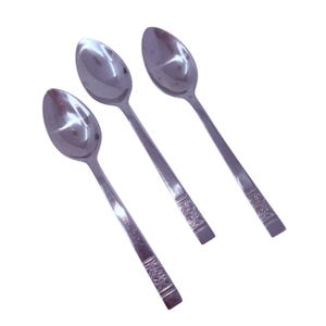May include: Three stainless steel spoons with oval bowls and long, slender handles. The handles have a decorative floral pattern near the base. The spoons are arranged diagonally against a white background.