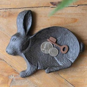 May include: A dark gray cast iron rabbit-shaped tray with a textured fur finish. Resting inside the tray are two silver-colored United States coins, a quarter and a dime, alongside a small, rusted brown metal skeleton key with a circular bow.