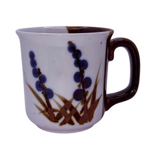 Puede incluir: Taza de cerámica blanca con asa y borde marrón. La taza presenta un diseño floral con bayas azules y tallos marrones. La taza es de tamaño y forma estándar, adecuada para bebidas calientes.