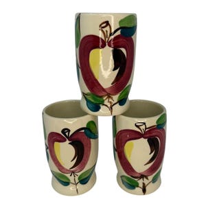 May include: Set of three white ceramic mugs with a hand-painted apple design. The apples are red, yellow, and brown with green leaves.