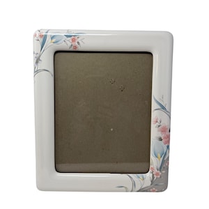 Könnte beinhalten: Ein weißer Keramik-Bilderrahmen mit rechteckiger Öffnung. Der Rahmen ist mit einem Blumenmuster mit rosa und blauen Blumen und grünen Blättern verziert. Der Rahmen hat eine glänzende Oberfläche.