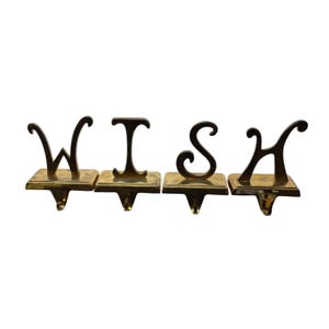 Puede incluir: Cuatro sujetadores de medias de latón que deletrean la palabra "WISH". Cada letra está montada sobre una base cuadrada con un gancho para colgar. Las letras tienen un estilo decorativo y cursivo.