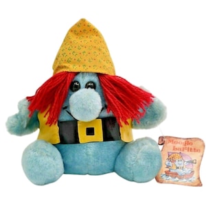 May include: A soft toy of a gnome-like figure with a blue body, red hair, and a yellow hat with a floral pattern. The gnome wears a yellow vest and a black belt with a gold buckle. A tag reads "Moogle la Fitte".
