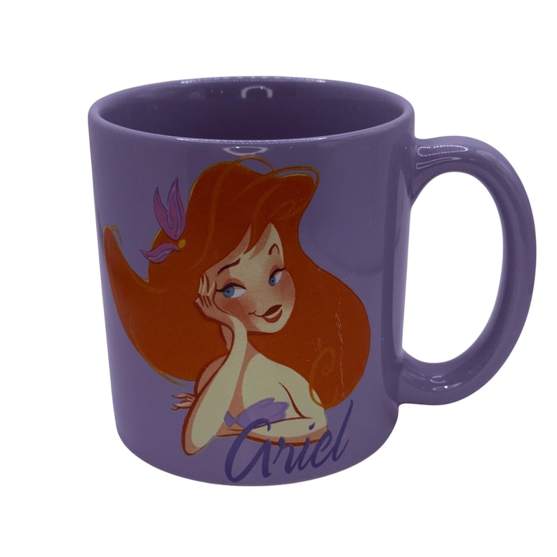Puede incluir: Una taza de cer&aacute;mica lila con una ilustraci&oacute;n de dibujos animados de Ariel de La Sirenita. Ariel tiene el pelo rojo, ojos azules y lleva un top morado. La taza tiene un asa curva y el nombre "Ariel" est&aacute; escrito en cursiva.