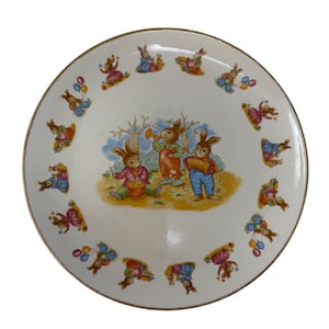 Pode incluir: Um prato de cerâmica branco com uma borda dourada apresenta uma cena caprichosa de três coelhos tocando música em uma floresta. O prato é decorado com uma borda de coelhos que realizam diversas atividades, incluindo brincar com balões.