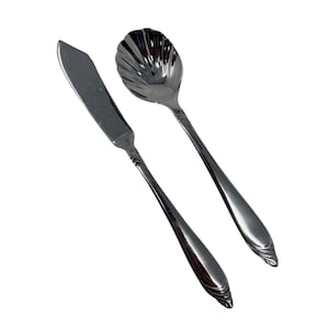 May include: A butter knife and a serving spoon set. The butter knife has a flat, rectangular blade and a decorative handle. The serving spoon has a shell-shaped bowl and a matching handle. Both are made of shiny, silver-colored metal.