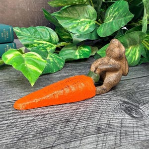 May include: A brown, textured rabbit figurine sitting upright and holding a large, bright orange carrot. The rabbit has a weathered, rustic appearance with visible ears and facial features, while the carrot has a tapered, ridged surface.