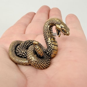 May include: A small, metallic gold-colored snake figurine with a coiled body and raised head. The surface features detailed, textured scales across the entire body, with a smooth underside on the neck and a slightly open mouth showing fangs.