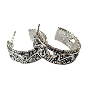 May include: A pair of silver-toned hoop earrings. Each earring features an intricate filigree design with a series of small, beaded accents along the edges. The earrings have a post closure.