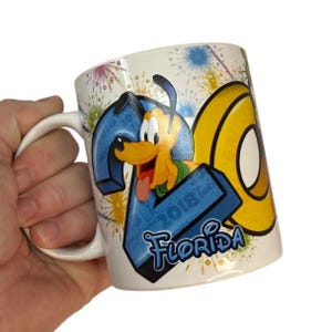 Peut inclure: Une tasse en céramique blanche avec un motif coloré représentant Pluto, des feux d'artifice et le texte "2018 Floride".