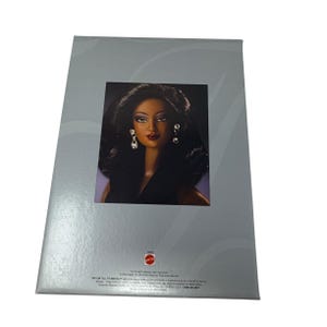 May include: A box with a picture of a doll. The doll has dark skin, long black hair, and is wearing earrings. The box is gray with a silver design and the Mattel logo.