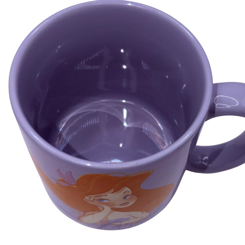 Puede incluir: Una taza de cer&aacute;mica lila con una ilustraci&oacute;n de sirena de dibujos animados. La sirena tiene el pelo rojo y un top morado. La taza tiene un asa curva y un acabado brillante. La taza es de tama&ntilde;o est&aacute;ndar.