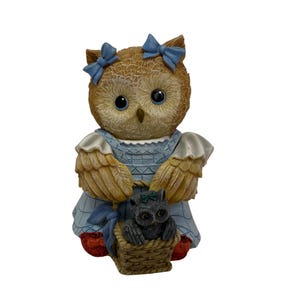 Puede incluir: Una figurita de búho marrón y azul que lleva un vestido azul y lleva una cesta con dos búhos pequeños dentro. El búho tiene lazos azules en la cabeza y ojos grandes y redondos.