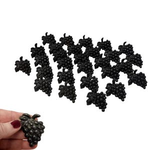 May include: A collection of black grape-shaped decorative elements. The elements are made of a dark material and feature detailed grape clusters and leaves. One element is held in a hand with red nail polish.
