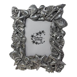 May include: A silver-toned picture frame with a detailed design of seashells and starfish. The frame has a rectangular opening and features the text "Sakura Vintage".