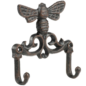 May include: A dark brown, textured metal wall hook featuring a bee motif at the top center. The design includes two curved hooks extending from a decorative, scroll-patterned base with two circular screw holes for mounting to a flat surface.