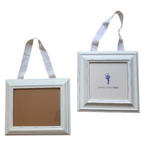 May include: Two white wooden picture frames with distressed edges and white ribbon hangers. One frame contains a brown backing, while the other features a white insert with the blue Pottery Barn Kids logo and a graphic of a child holding a frame.