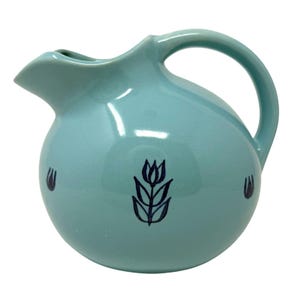 Può includere: Una brocca in ceramica turchese con corpo arrotondato e manico curvo. La brocca ha un beccuccio per versare ed è decorata con disegni floreali blu scuro. Il design è semplice ed elegante, adatto per servire bevande.