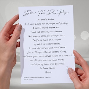 May include: A white sheet of paper featuring the text "Daniel Fast Deep Prayer" in a decorative script. The prayer, printed in black, is held by hands with manicured nails. The text includes a reference to Daniel 10:12.