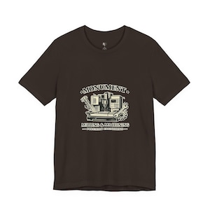 Pode incluir: Camiseta marrom escuro com um design gráfico. O design apresenta o texto "MONUMENT" acima de "MILLING & MACHINING" e "PRECISION ENGINEERING". O gráfico inclui uma fresadora e ferramentas.