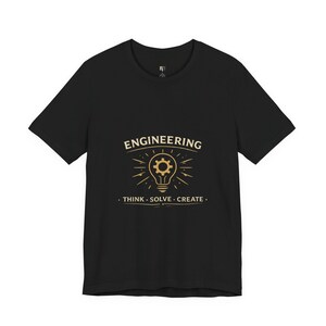 Puede incluir: Camiseta negra con un gráfico dorado. El gráfico presenta la palabra "ENGINEERING" encima de un diseño de bombilla con un engranaje en su interior. Debajo, el texto dice "THINK . SOLVE . CREATE."