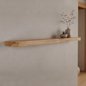 May include: A light-colored wooden floating shelf mounted on a neutral-toned wall. The shelf holds a small vase with a plant and a small decorative pot. The shelf is approximately 40 inches long.