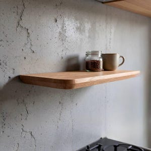 以下が含まれることがあります： 灰色のコンクリート壁に取り付けられた、丸みを帯びた前面エッジを持つフローティング木製棚。茶色の豆が入ったガラス瓶とベージュのマグカップが棚の上に置かれています。棚は明るい色の木材で作られています。
