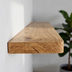 May include: A rectangular floating wall shelf made of natural light brown wood with a visible grain pattern, a prominent knot, and a smooth, rounded front edge. The shelf is mounted horizontally against a plain, light-coloured wall surface.