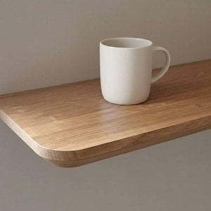 May include: A light-colored wooden wall shelf with rounded corners and a smooth grain pattern. A plain, matte off-white ceramic mug with a curved handle sits on the surface of the shelf. The shelf is mounted horizontally against a neutral wall.