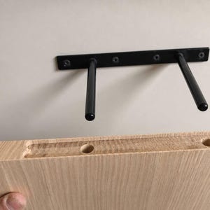 K&ouml;nnte beinhalten: Ein schwarzer Metall-Regaltr&auml;ger f&uuml;r schwebende Regale mit zwei horizontalen Stangen an einer Wand, positioniert &uuml;ber einem hellen Holzregalbrett mit zwei vorgebohrten kreisf&ouml;rmigen L&ouml;chern und einer Nut, die f&uuml;r die Stangen ausgelegt ist.