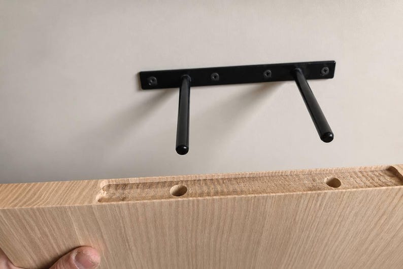 K&ouml;nnte beinhalten: Ein schwarzer Metall-Regaltr&auml;ger f&uuml;r schwebende Regale mit zwei horizontalen Stangen an einer Wand, positioniert &uuml;ber einem hellen Holzregalbrett mit zwei vorgebohrten kreisf&ouml;rmigen L&ouml;chern und einer Nut, die f&uuml;r die Stangen ausgelegt ist.