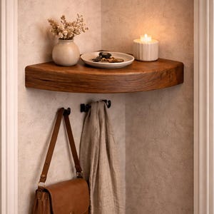 May include: A wooden corner shelf mounted on a wall, featuring a curved front edge. Below the shelf are two black metal hooks holding a brown leather shoulder bag and a beige fabric scarf. On the shelf sits a small vase, a dish, and a candle.