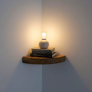K&ouml;nnte beinhalten: Ein geschwungenes Eckregal aus Holz, das an zwei W&auml;nden montiert ist. Auf dem Regal liegt ein Stapel aus drei Hardcover-B&uuml;chern, gekr&ouml;nt von einer kleinen, runden, wei&szlig;en Keramiklampe, die ein warmes Licht in die Ecke abgibt.