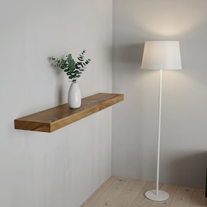 May include: A rectangular wooden floating shelf mounted on a wall, holding a white ceramic vase with green eucalyptus stems. Beside the shelf stands a tall, thin white floor lamp with a white drum shade that emits a soft, warm light.