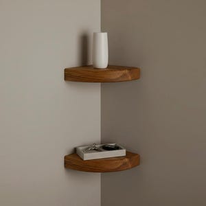 May include: Two floating corner shelves made of natural wood with a visible grain pattern. The top shelf holds a tall, smooth white cylindrical vase. The bottom shelf holds a rectangular white tray containing a set of metal keys and a black fob.