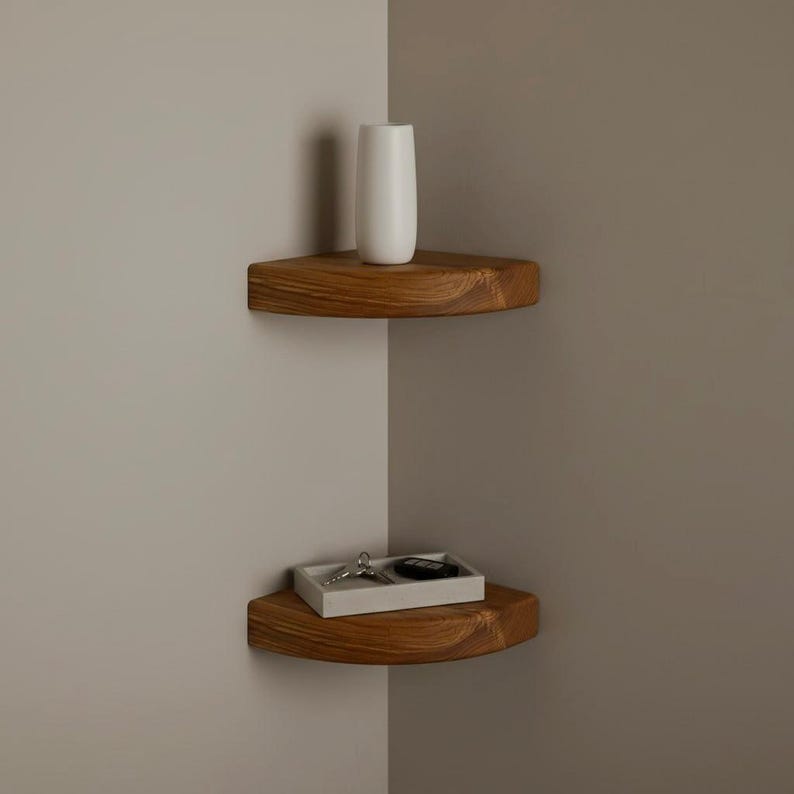 May include: Two floating corner shelves made of natural wood with a visible grain pattern. The top shelf holds a tall, smooth white cylindrical vase. The bottom shelf holds a rectangular white tray containing a set of metal keys and a black fob.