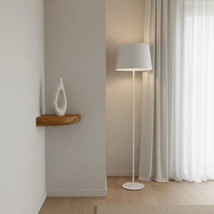 K&ouml;nnte beinhalten: Eine wei&szlig;e Stehlampe mit zylindrischem Schirm und d&uuml;nnem Metallst&auml;nder, die neben einer Wandecke steht. Ein kleines, geschwungenes Eckregal aus Holz ist an der Wand montiert und tr&auml;gt eine glatte, wei&szlig;e, abstrakte Keramikskulptur mit einer &Ouml;ffnung.