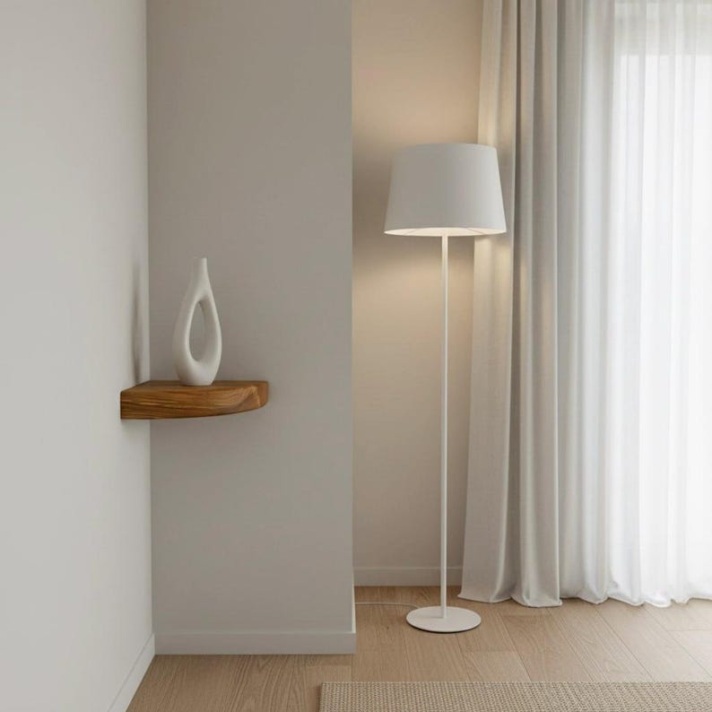 K&ouml;nnte beinhalten: Eine wei&szlig;e Stehlampe mit zylindrischem Schirm und d&uuml;nnem Metallst&auml;nder, die neben einer Wandecke steht. Ein kleines, geschwungenes Eckregal aus Holz ist an der Wand montiert und tr&auml;gt eine glatte, wei&szlig;e, abstrakte Keramikskulptur mit einer &Ouml;ffnung.