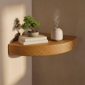 K&ouml;nnte beinhalten: Ein geschwungenes Eckregal aus Holz an einer Wand. Auf dem Regal steht ein kleiner gr&uuml;ner Bonsai in einem runden grauen Topf auf zwei gestapelten wei&szlig;en B&uuml;chern sowie ein glatter, cremewei&szlig;er, abgerundeter Diffusor, der einen feinen Dampf abgibt.