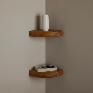 May include: Two wooden corner shelves. The top shelf holds a white vase, and the bottom shelf has a small tray with keys and a key fob. The shelves are made of natural wood and are mounted in a corner.