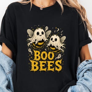 May include: A black t-shirt featuring a graphic of two bees wearing white ghost costumes. The bees have yellow and black striped bodies and small wings. The text "BOO BEES" is printed in a distressed yellow font below the bees, surrounded by small ghost and star illustrations.