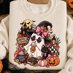 May include: A cream-colored sweatshirt featuring a graphic of a ghost wearing a witch hat with a pink bow and heart-shaped sunglasses, holding a drink. The design includes a black cat, pumpkins, a haunted house, and text reading Spooky Season and Basic Witch.