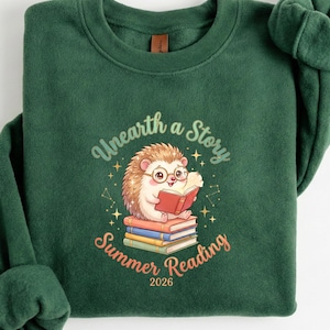 May include: A dark green sweatshirt with the text "Unearth a Story Summer Reading 2026." The graphic features a cartoon hedgehog wearing glasses and reading a book while sitting on a stack of books. The design includes stars and a constellation.