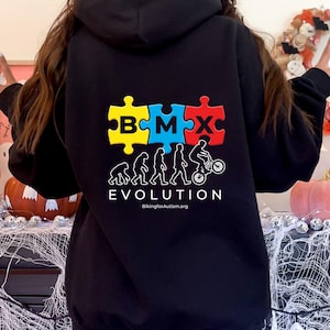 Può includere: Felpa con cappuccio nera con un design a puzzle colorato con le lettere BMX. Sotto, una grafica evolutiva di una persona che va in bicicletta BMX. La parola "EVOLUTION" è stampata sotto, con il sito web "BikingforAutism.org".