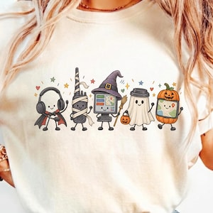 Könnte beinhalten: Ein cremefarbenes T-Shirt mit einer Grafik von fünf Zeichentrickfiguren in Halloween-Kostümen: ein Vampir mit Kopfhörern, ein Funkgerät als Mumie, ein Computer mit Hexenhut, ein Geist mit Kürbiseimer und ein GPS-Gerät als Kürbis.